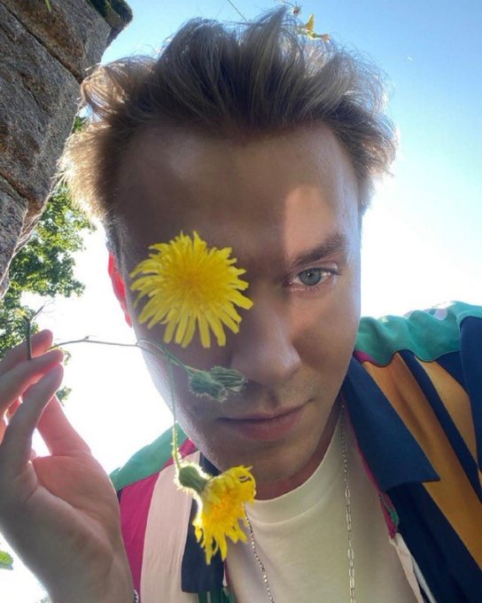 A young man taking a selfie outdoors, holding a yellow dandelion that covers one of his eyes, wearing a colorful striped shirt and silver chain.