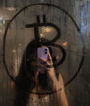 In this image, a woman is taking a selfie using her cell phone that features a Bitcoin symbol drawn or stenciled over the photo of herself. The context behind her decision to include a Bitcoin-related image might be an interest or affiliation with the cryptocurrency world, although it's not explicitly clear from the image alone.