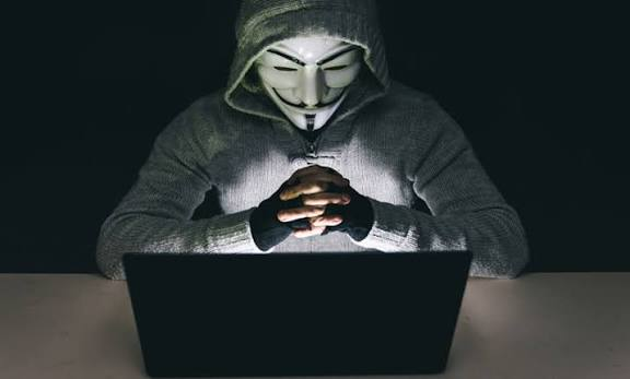 A figure wearing a Guy Fawkes mask and a hooded gray sweater sits in darkness, illuminated only by the glow of a laptop screen with hands clasped.