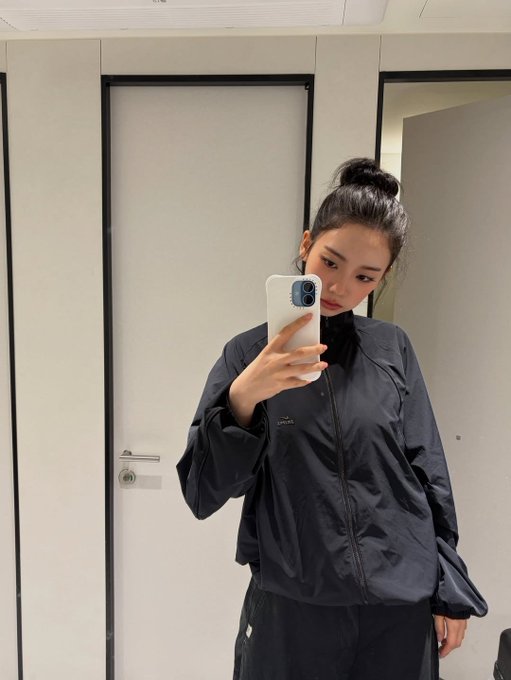 A young woman takes a mirror selfie wearing a black Lacoste windbreaker, her hair pulled up in a high bun. The "LACOSTE" text and crocodile logo are visible on the jacket, perfectly capturing the casual "Mirror" selfie vibe.