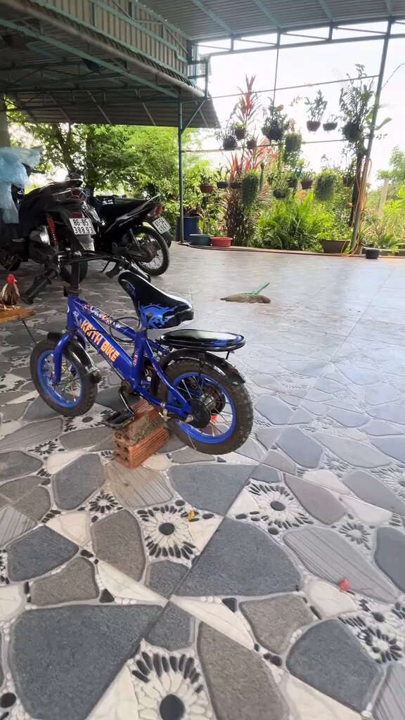 The image features a blue bicycle parked next to a building, with a tiled floor beneath it. The bicycle has a wooden stand attached to its back wheel, providing support for the bike's front tire. In the background, there are several potted plants scattered around the area, adding greenery and life to the scene. A motorcycle is also visible in the image, parked nearby.