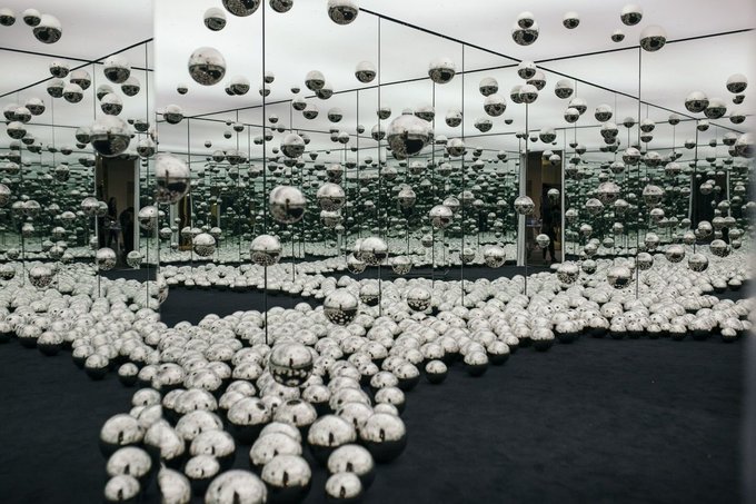 An art installation featuring countless reflective silver spheres of various sizes scattered on a dark floor and suspended in a mirrored room, creating infinite reflections.