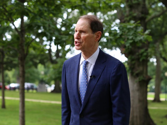 Senator Ron Wyden, wearing a navy blue suit and blue patterned tie, speaks to reporters outdoors in a tree-lined setting. His advocacy in the accompanying post—pushing to preserve Section 604 of the CLARITY Act—relates to his ongoing role shaping cryptocurrency and blockchain policy in the Senate.