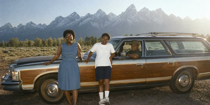A vintage photograph showing three people—a woman in a blue dress, a young man in a white shirt, and a man in a station wagon—posed in front of wood-paneled car with snow-capped mountains in the background.
