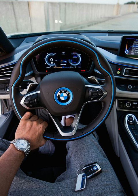 A person's hand, adorned with a luxury watch, grips a BMW steering wheel featuring the iconic blue-accented logo, with the car's digital dashboard and infotainment screen visible in the background, while a BMW key fob rests on their lap—picturing a luxurious ride that aligns with the post's "money making Monday" theme of seeking blessings and new opportunities.