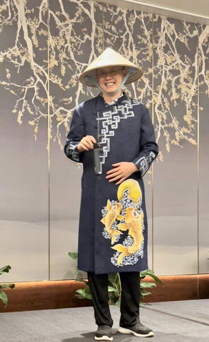 A smiling young person poses wearing a traditional Vietnamese áo dài with blue dragon and wave designs, a conical nón lá hat, and black pants, set against a backdrop of stylized golden branches. The image accompanies a greeting in Vietnamese ("Xin chào" meaning "Hello") directed at CZ Binance, the founder of the cryptocurrency exchange Binance.