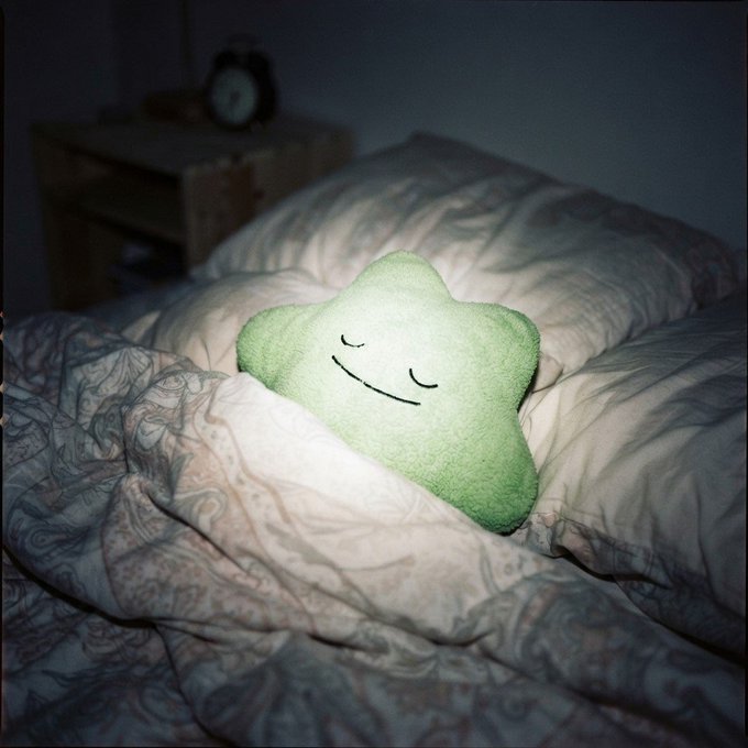 A green star-shaped plush pillow with closed eyes rests on a rumpled bed with patterned bedding in a dimly lit bedroom.