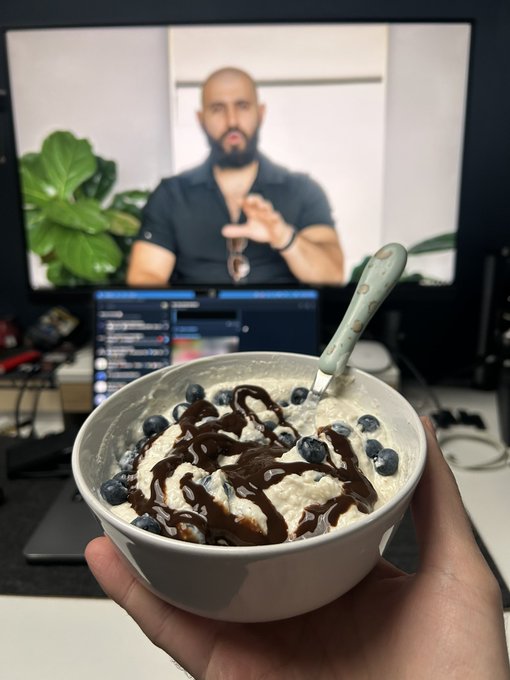 A hand holds a bowl of ice cream topped with blueberries and chocolate syrup in front of a computer monitor displaying a man with a beard on a video call.