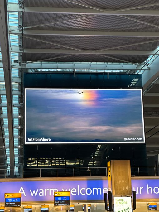 An airport terminal displaying a large screen showing an aerial photograph of a bird in flight with rainbow colors, labeled 'ArtFromAbove @artorush.com', with 'A warm welcome' text visible below.