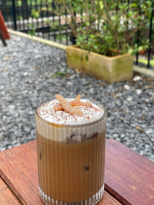 An iced coffee drink topped with foam, cocoa powder, and small pastry pieces served in a ridged glass on a wooden table, set in an outdoor cafe area with plants in the background.