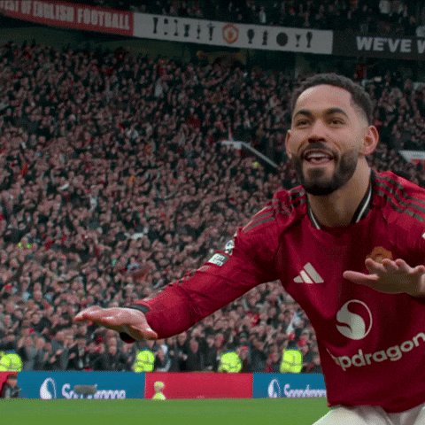 The image shows a man wearing a red soccer jersey, celebrating his goal with both arms outstretched. He appears to be in a stadium filled with fans, as evidenced by the crowd of people visible in the background. The scene captures the excitement and joy of the moment, with the man's body language conveying a sense of triumph and satisfaction.