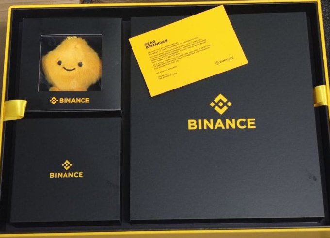 An open Binance gift box containing a fluffy yellow plush character in a display window, a smaller black box with the Binance logo, and a yellow 'DEAR BINANCIAN' letter on top of a larger black Binance-branded package.