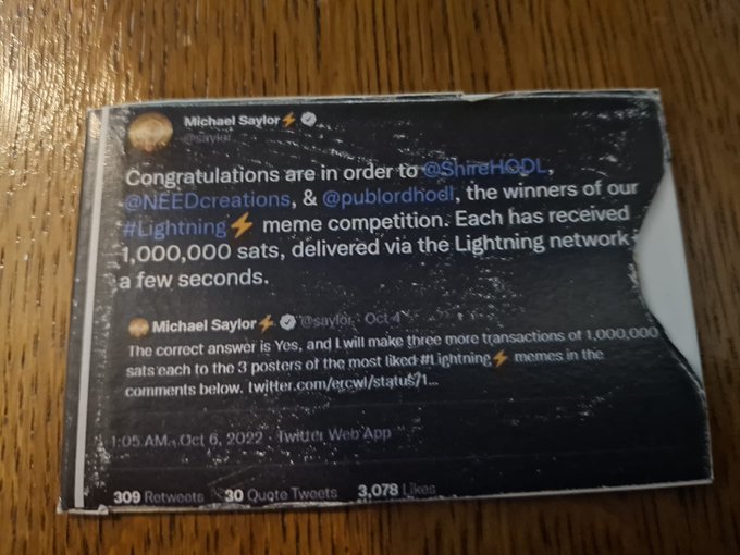 A worn photograph shows a tweet screenshot from Michael Saylor (@saylor) announcing winners of a Lightning meme competition who each received 1,000,000 sats, with 309 retweets, 30 quote tweets, and 3,078 likes dated October 6, 2022—serving as proof of the Bitcoin tip referenced in the caption.