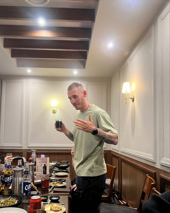 A man in a light green t-shirt stands in an elegant restaurant with wood-beamed ceilings, holding a glass of dark beverage and gesturing with his hand while a table full of dishes and Cass beer bottles sits before him.