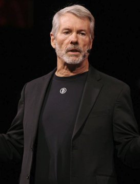 Michael Saylor, founder of Strategy (formerly MicroStrategy), speaks on stage wearing a black blazer over a t-shirt featuring a small "B" logo. The image illustrates the post's commentary on how Saylor, once idolized by the Bitcoin community for his aggressive corporate BTC purchases, is now reportedly considering selling or laying the groundwork to do so.