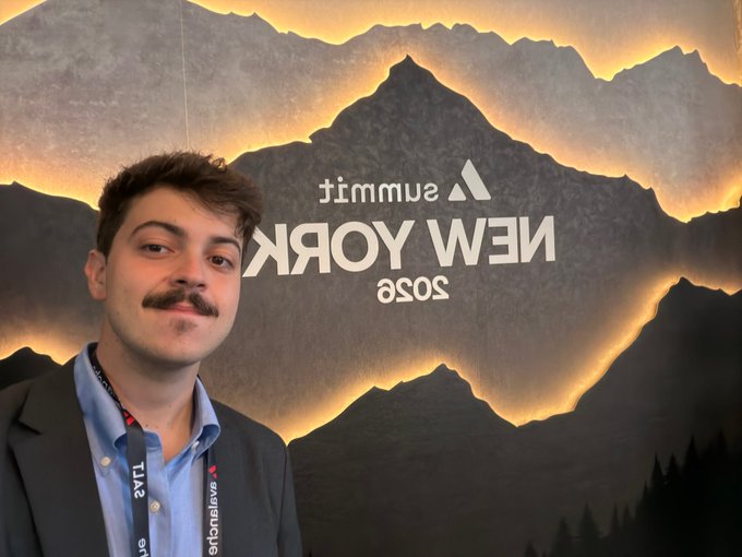 A young man with a mustache poses in front of a 'Summit New York 2025' backdrop featuring a mountain graphic.