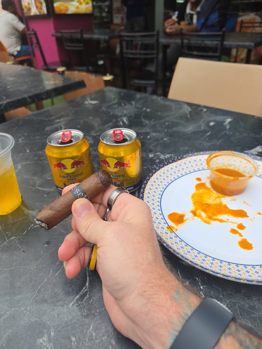 A hand holds a half-smoked cigar alongside two cans of Red Bull on a marble table, with a plate of hot sauce smeared around a small cup. The image captures the post-conference vibe of @Token2049, where "breakfast of degens" refers to fueling late-night crypto networking with energy drinks and cigars.