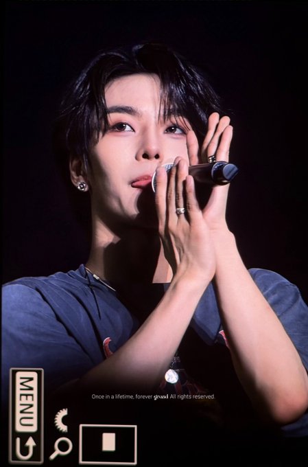 The image shows a young man with dark hair performing on stage, holding a microphone close to his face with both hands, wearing a blue t-shirt and rings on his fingers. The post's hashtags identify him as Xinlong of ALPHADRIVE ONE, and the caption expresses how seeing fans happy brings him joy, reflected in his warm, engaged expression toward the audience. Visible text includes "MENU," "Once in a lifetime, forever grateful. All rights reserved," and the date "260626."