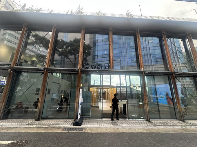 The image shows the exterior of a modern, glass-fronted "world" branded building (Worldcoin's iris registration center) in what appears to be a Korean urban area, with a person standing at the entrance. The caption and post confirm this is Worldcoin's surprisingly large registration facility in Ikseon-dong, Korea, with the author noting their surprise at the scale despite Korea no longer offering iris scan compensation.