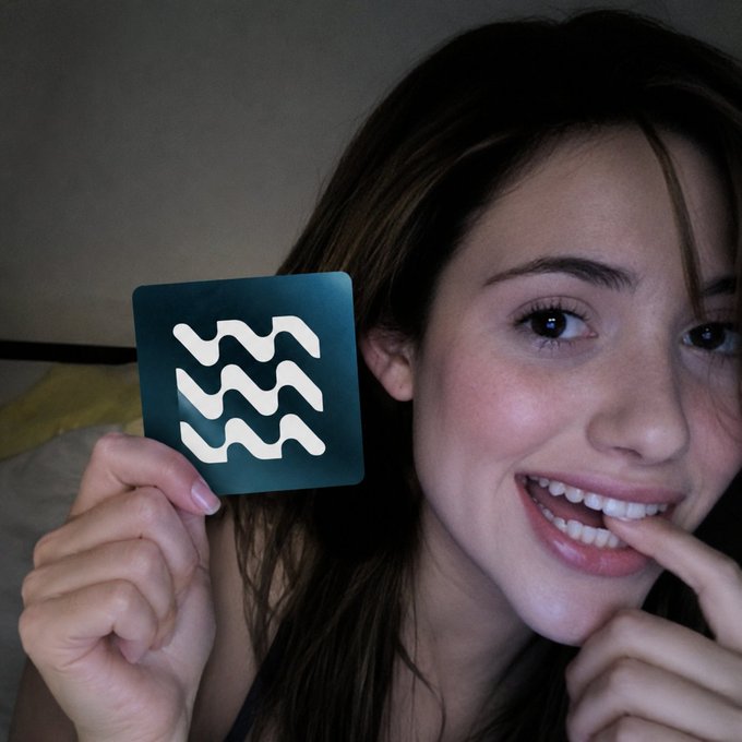 A smiling young woman holds up a dark square icon featuring a white wavy water symbol (the Aquarius zodiac sign) next to her face.
