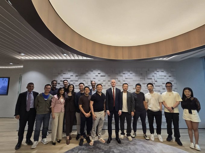 A group of approximately twenty people pose together for a formal photo in a modern office space featuring a distinctive circular light fixture and textured wall.