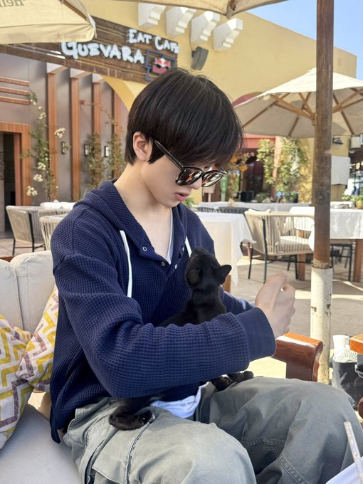 A digitally altered photo of a young man wearing sunglasses and a blue knit hoodie sitting at an outdoor café called 'EAT CAFE QUEVARA,' holding a small black kitten in his lap.