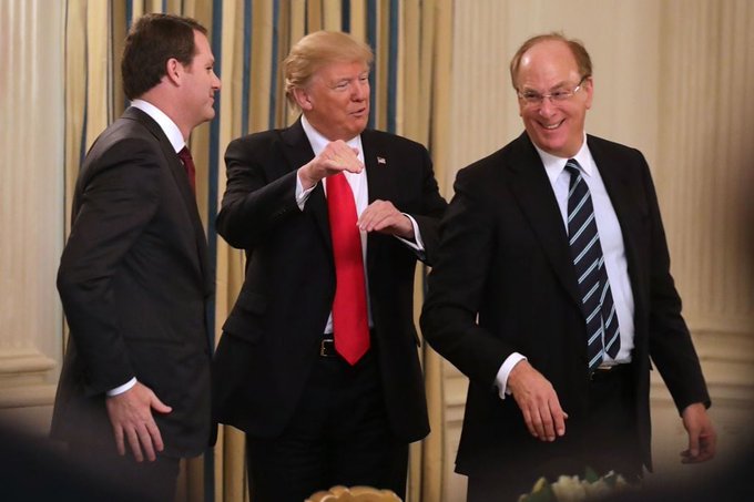 The image shows three men dressed in suits standing next to each other against a white wall with blue stripes. One of them is wearing glasses and pointing at something outside the frame. The man in the center is holding up his hand, possibly gesturing or making a point. In the background, there are two more people partially visible on the right side of the image.