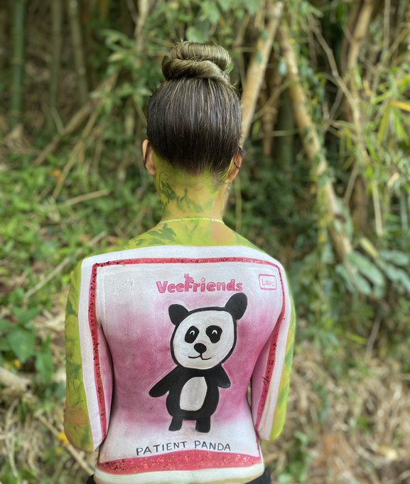 Body art on a person's back featuring a 'VeeFriends Patient Panda' trading card design and green leaf patterns on their shoulders, set against a bamboo background.