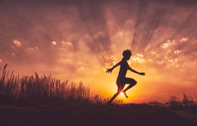 A silhouette of a person joyfully jumping against a dramatic orange sunset sky with rays of light bursting through the clouds.