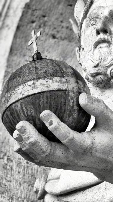 A black-and-white close-up photograph of a weathered stone statue of a bearded man holding an orb topped with a cross.