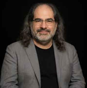 The image features a man with long hair wearing glasses, smiling at the camera while sitting down. The background of the photo is black, which contrasts with the man's appearance. There are no texts or other objects present in the image.