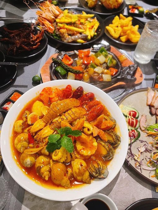 A table spread featuring a large bowl of seafood stew with lobster, abalone, and various vegetables, surrounded by other dishes including prawns, fries, and a salad.