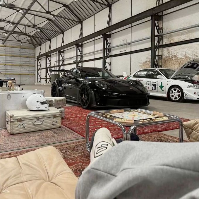A view inside a spacious garage/warehouse showing a black Porsche sports car with a large rear wing and a white race car parked on a concrete floor, with vintage suitcases, a white helmet, and a glass coffee table in the foreground.