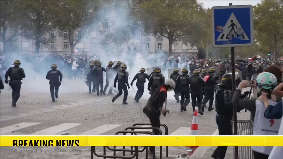 A breaking news screenshot showing riot police in helmets charging through tear gas during a street protest, with a pedestrian crossing sign and barricades visible.
