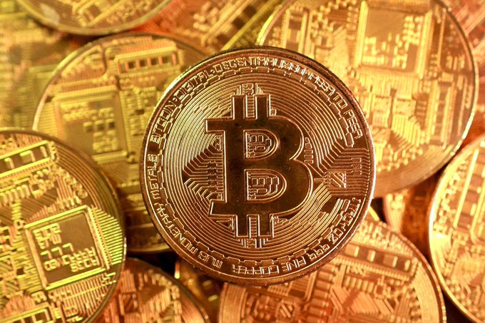A close-up image of multiple gold-colored Bitcoin (BTC) physical tokens or commemorative coins, with the iconic Bitcoin 'B' symbol prominently displayed on the central coin surrounded by circuitry-like engravings.