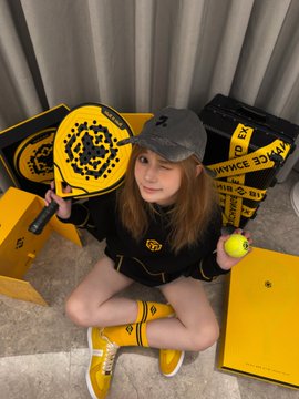 A young woman sits on the floor wearing a black and yellow outfit, holding a matching yellow tennis racket with a smiley face design and a tennis ball, surrounded by yellow and black boxes.