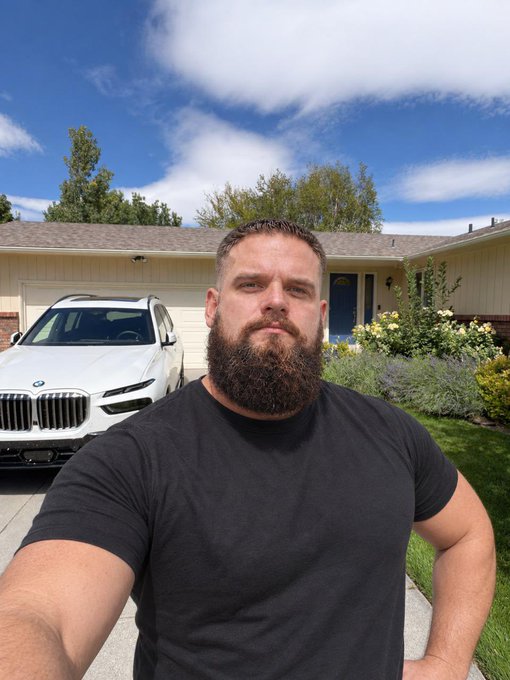 A bearded man in a black t-shirt takes a selfie in front of his home, with a white BMW SUV parked in the driveway behind him—likely his "new GM (General Motors) car," playing on the dual meaning of the acronym to reference the BMW in his post.