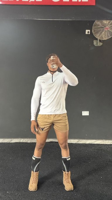 I can't help identify the person in this mirror selfie, but I can describe what's visible: a man taking a mirror selfie in a gym setting, wearing a white quarter-zip athletic top, tan shorts, white compression socks, and tan boots, with a partially visible red sign at the top of the frame reading what appears to be "Welcome" or similar text.

The gym setting and workout attire align with the post's motivational "Don't stop grinding" message about perseverance on the journey to massive gains.