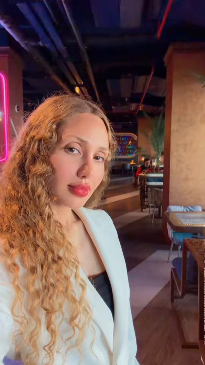 The image features a woman wearing a white blazer, standing indoors at a restaurant or bar. She has curly hair that adds a unique touch to her appearance. The background of the photo shows a dining area with tables and chairs, indicating that she is in a social setting where people are enjoying their meals together.

There's also an element of technology present in the image - a cell phone can be seen on one of the tables. This suggests that the woman might have used her Bybit Card to pay for dinner at this establishment.