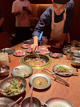 A chef in a blue shirt and apron cooking meat on a tabletop Korean BBQ grill, surrounded by various plates of raw meat, side dishes, and sauces.