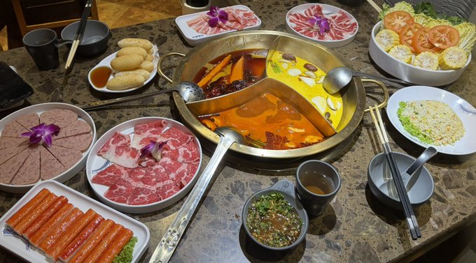 A table full of hot pot ingredients including a divided pot of spicy and mild broth, sliced meats, crab sticks, tomatoes, tofu, spring rolls, and various dipping sauces.