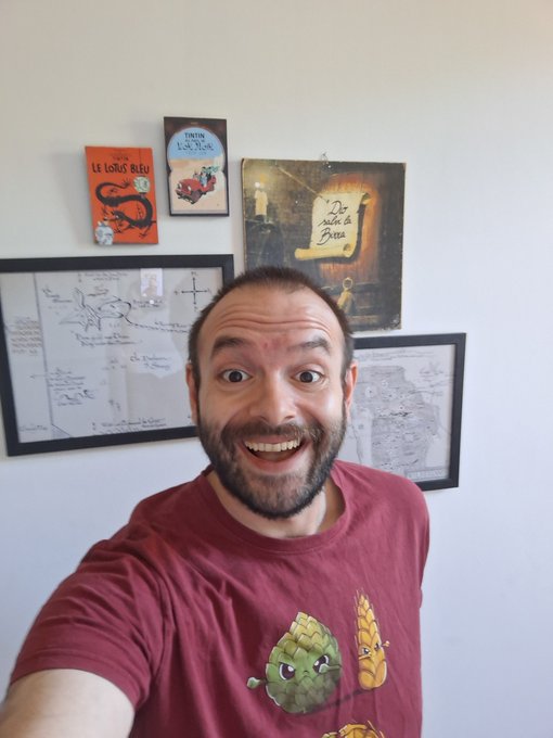 The image features a man wearing a red shirt, standing against a white wall with several framed pictures hanging above him. The man has a big smile on his face as he takes a selfie using his phone's camera. The caption of the photo reads "Gooooood morning beautieeeees! -Video about Elowyn in the next post -Stop wasting time chatting with Greenity -Say Something at 1 p.m. - Edit the next video -Space in Italian tonight with Panofsky and the OOBE PROTOCOL team Lots to do today!".