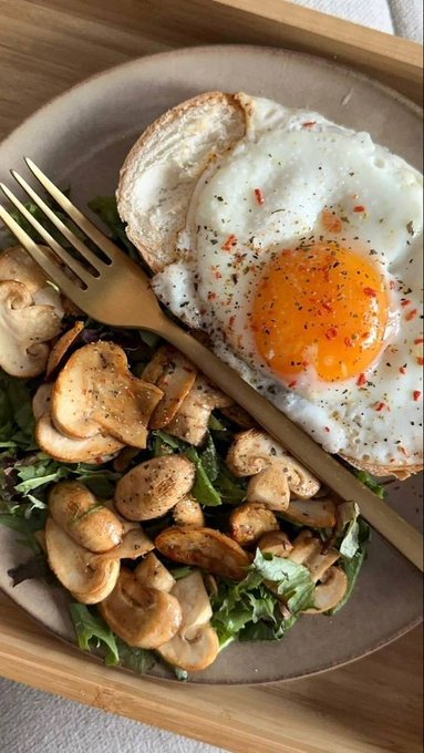 An overhead photo of a meal on a beige plate: a fried egg with a runny yolk and red seasoning sprinkled on top, served over toast and accompanied by sautéed mushrooms and greens, with a gold fork resting on the plate.