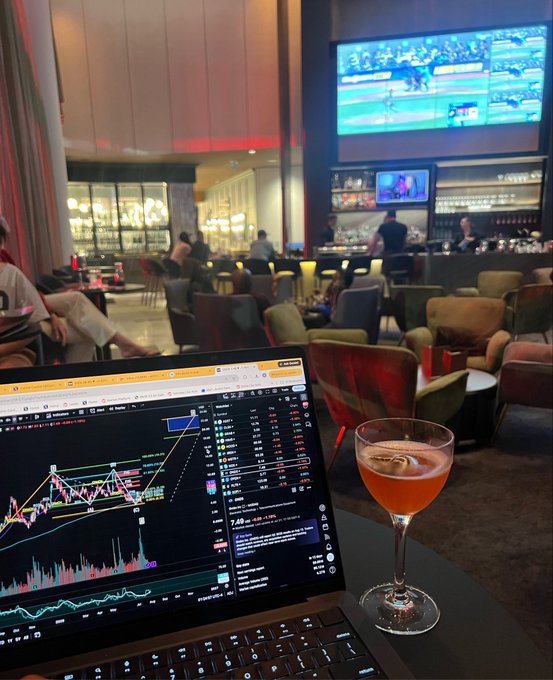 A person works on a laptop displaying a $ONDS (Ondas Holdings) stock chart showing the price at $7.49, accompanied by a cocktail glass, capturing a bullish Saturday night trading session in what appears to be a hotel lounge or sports bar with multiple TVs on the wall.