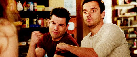 Two actors, dressed casually and with serious expressions, sit at a table in a library or bookstore setting.