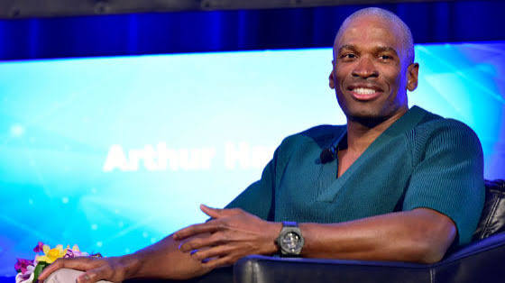 Arthur Hayes is seated on a stage, smiling, with his name displayed on a large screen behind him.