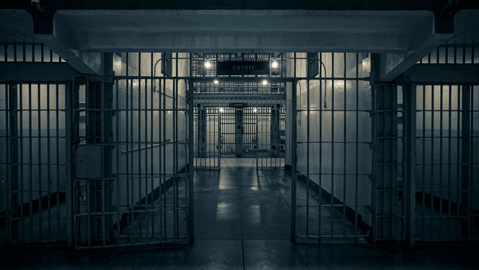 A dark, atmospheric view down a prison corridor with metal barred cells on either side and a sign reading 'CUTOFF' hanging from the ceiling.