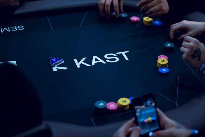 An overhead view of a poker table branded "KAST," showing players' hands and colorful stacks of chips along with a phone displaying the game.