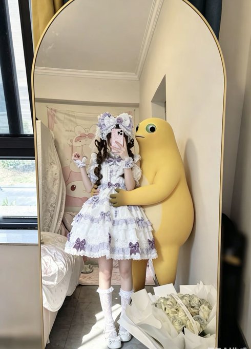 A person in a purple and white frilly Lolita dress takes a mirror selfie with a pink phone, standing next to a large yellow cartoon bear plushie and a bouquet of white roses.