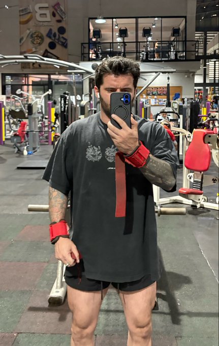 A man with a beard and tattoos takes a mirror selfie in a gym, wearing a dark gray t-shirt, black shorts, and red wrist wraps.